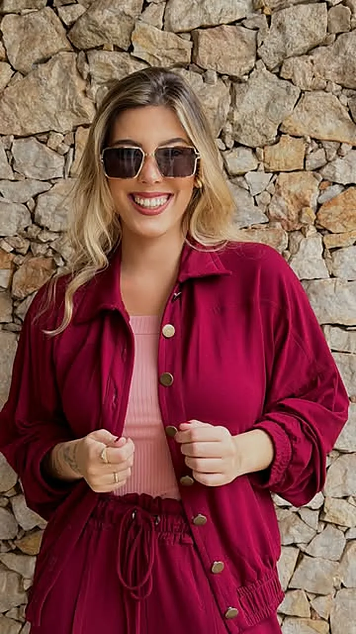 Mulher de conjunto vinho e óculos de sol diante de um muro de pedra