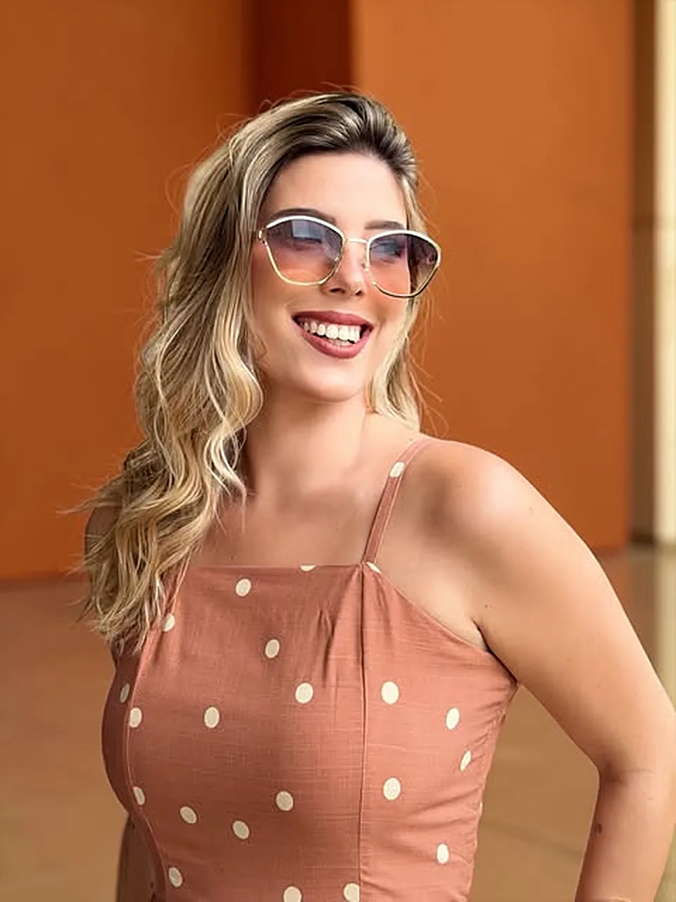 Mulher sorrindo com óculos de sol de armação clara, em frente a uma parede terracota